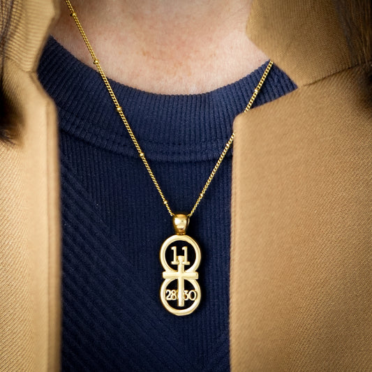 The RIYAN 29 and 11® pendant has the numbers 11, 28, and 30 intertwined with the cross to represent Matthew 11:28-30 with the chapter word "Matthew" inscribed on the back.  Model is wearing a large Riyan pendant on our 18" Satellite chain.  Model is wearing a navy high neck tank and a camel blazer.