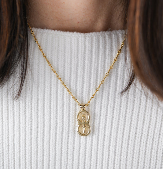 The RIYAN 29 and 11® pendant has the numbers 5 and 7 intertwined with the cross to represent 2 Corinthians 5:7 with the chapter name "2 Corinthians" inscribed on the back.  The model is wearing a large Riyan pendant on our 18" Rolo chain against a white ribbed sweater.