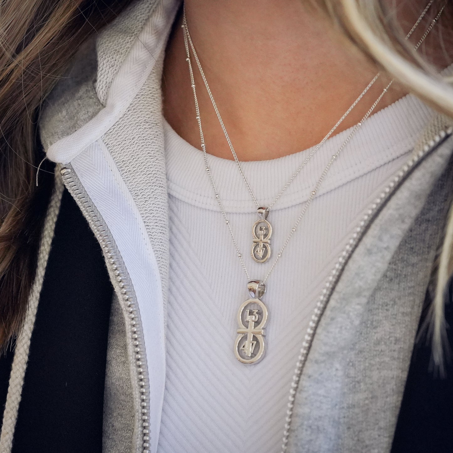 The silver RIYAN 29 and 11® pendant has the numbers 13, 4, and 4 intertwined with the cross to represent 1 Corinthians 13:4-7 with the chapter word "1 Corinthians" inscribed on the back.  Model is wearing the large silver pendant on an 18" Wheat chain and a small Riyan pendant on a 16" satellite chain.  Model is wearing a Veronica Beard blazer and grey hoodie sweatshirt.