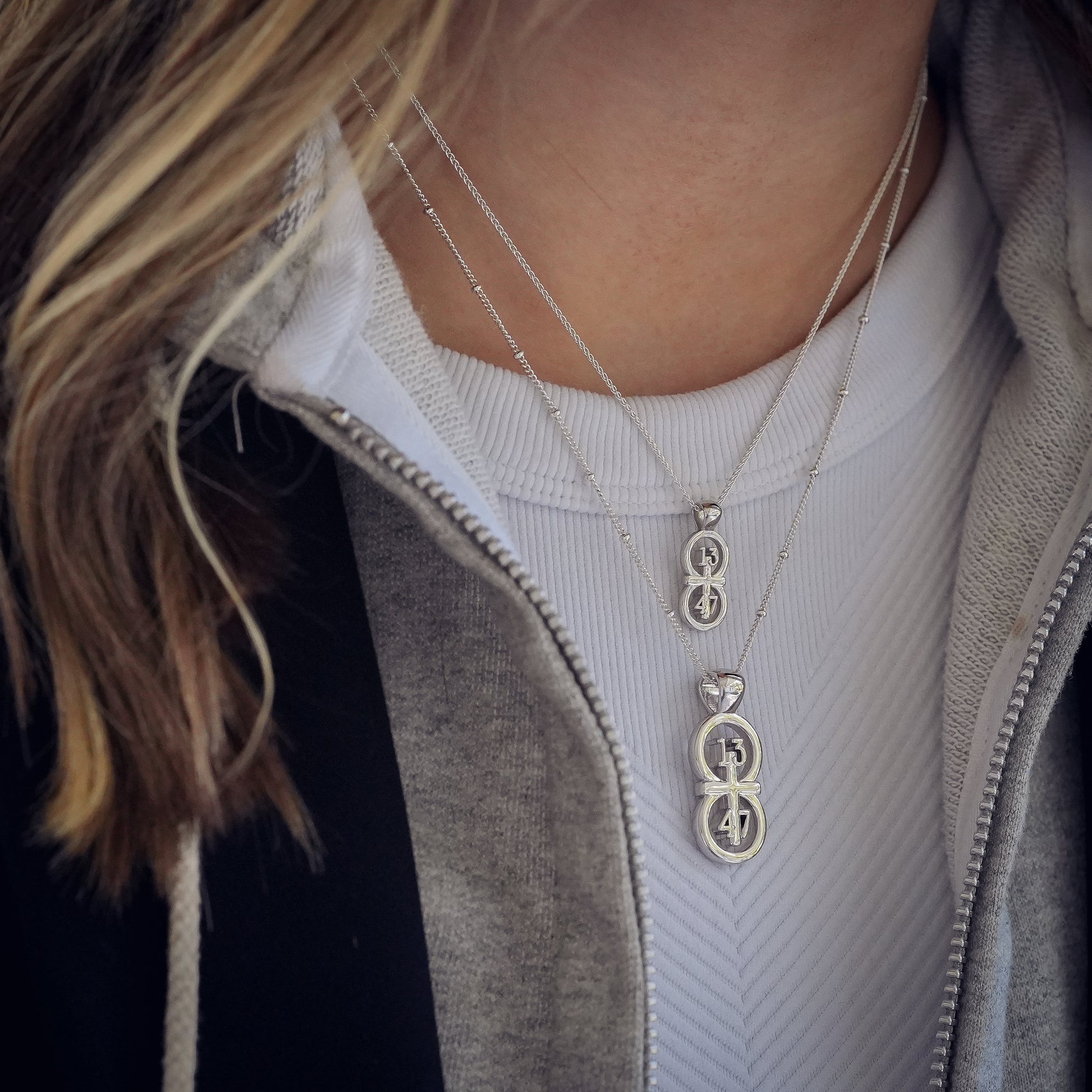 The silver RIYAN 29 and 11® pendant has the numbers 13, 4, and 4 intertwined with the cross to represent 1 Corinthians 13:4-7 with the chapter word "1 Corinthians" inscribed on the back.  Model is wearing the large silver pendant on an 18" Satellite chain and a small Riyan pendant on a 16" satellite chain.  Model is wearing a Veronica Beard blazer and grey hoodie