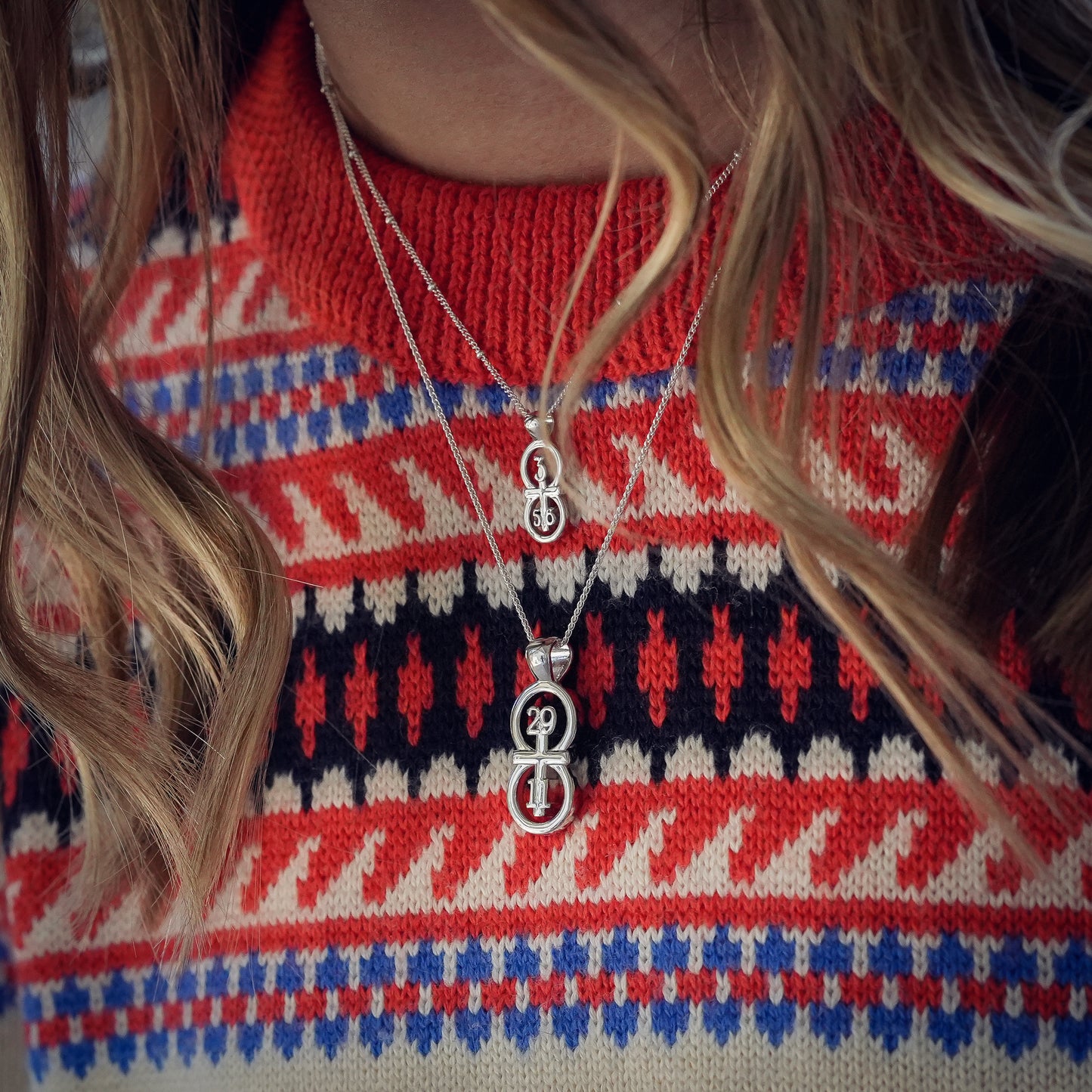 The RIYAN 29 and 11® silver pendant has the numbers 29 and 11 intertwined with the cross to represent Jeremiah 29:11 with the chapter word "Jeremiah" inscribed on the back. Model is wearing the large silver Riyan pendant on an 18" Wheat chain and a small silver Riyan pendant on a 16" Satellite chain. Model is wearing a high neck colorful sweater.