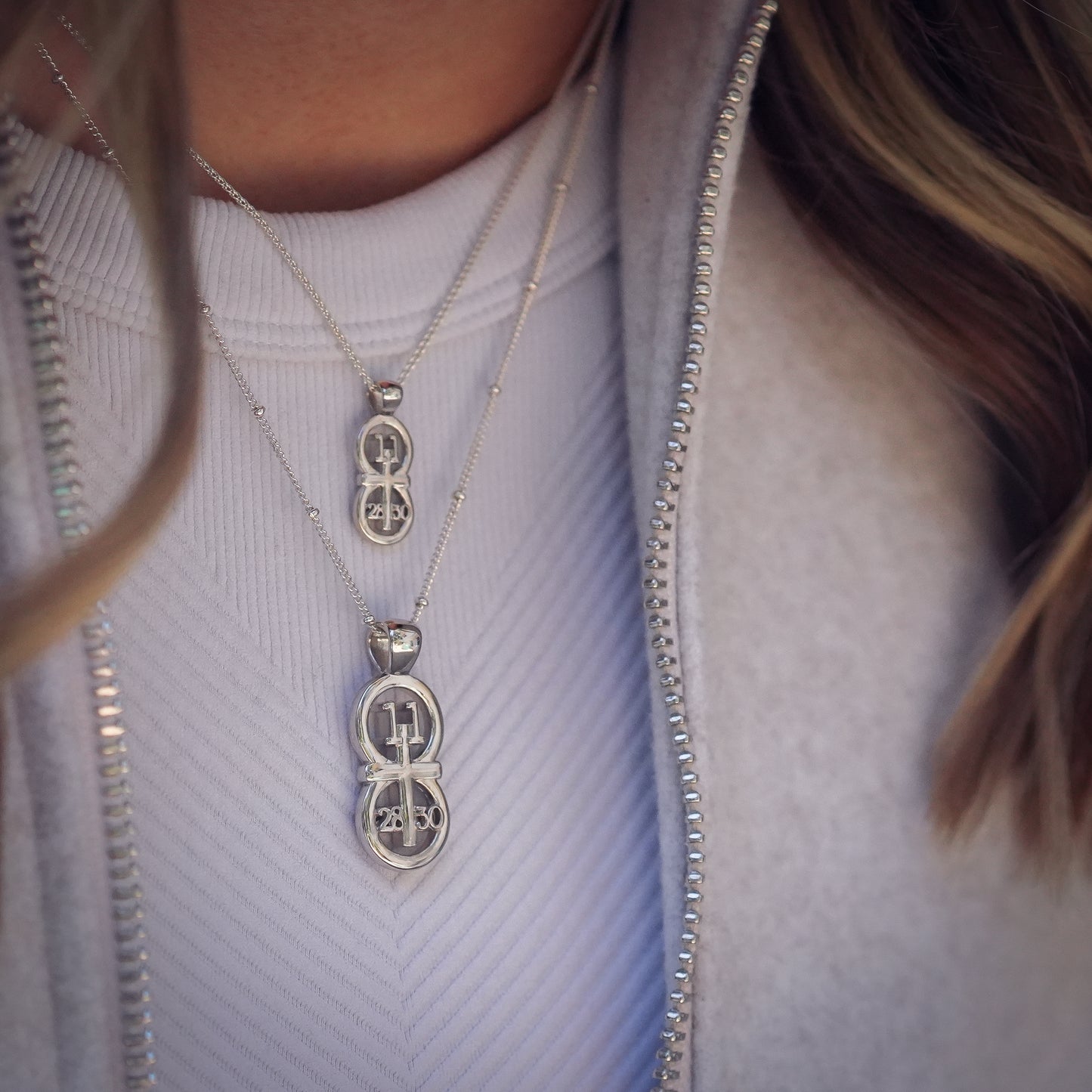 The RIYAN 29 and 11® silver pendant has the numbers 11, 28, and 30 intertwined with the cross to represent Matthew 11:28-30 with the chapter word "Matthew" inscribed on the back. The model is wearing a large Riyan pendant on our 18" Satellite Chain and a small silver Riyan pendant on our 16" Wheat chain. Model is wearing a white high neck tank and a grey wool jacket.