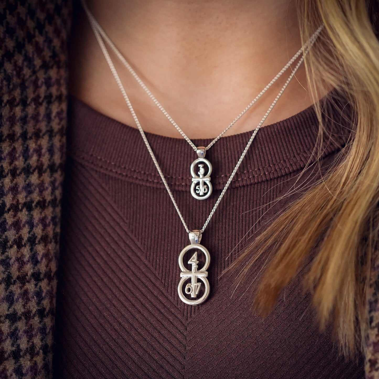 The silver RIYAN 29 and 11® pendant has the numbers 4,6, and 7 intertwined with the cross to represent Philippians 4:6-7 with the chapter name "Phillipians" inscribed on the back. Model is wearing a large silver Riyan pendant on our 18" Wheat chain and a small silver Riyan pendant on our 16" Wheat chain.  Model is wearing a burgundy tank and a houndstooth blazer.