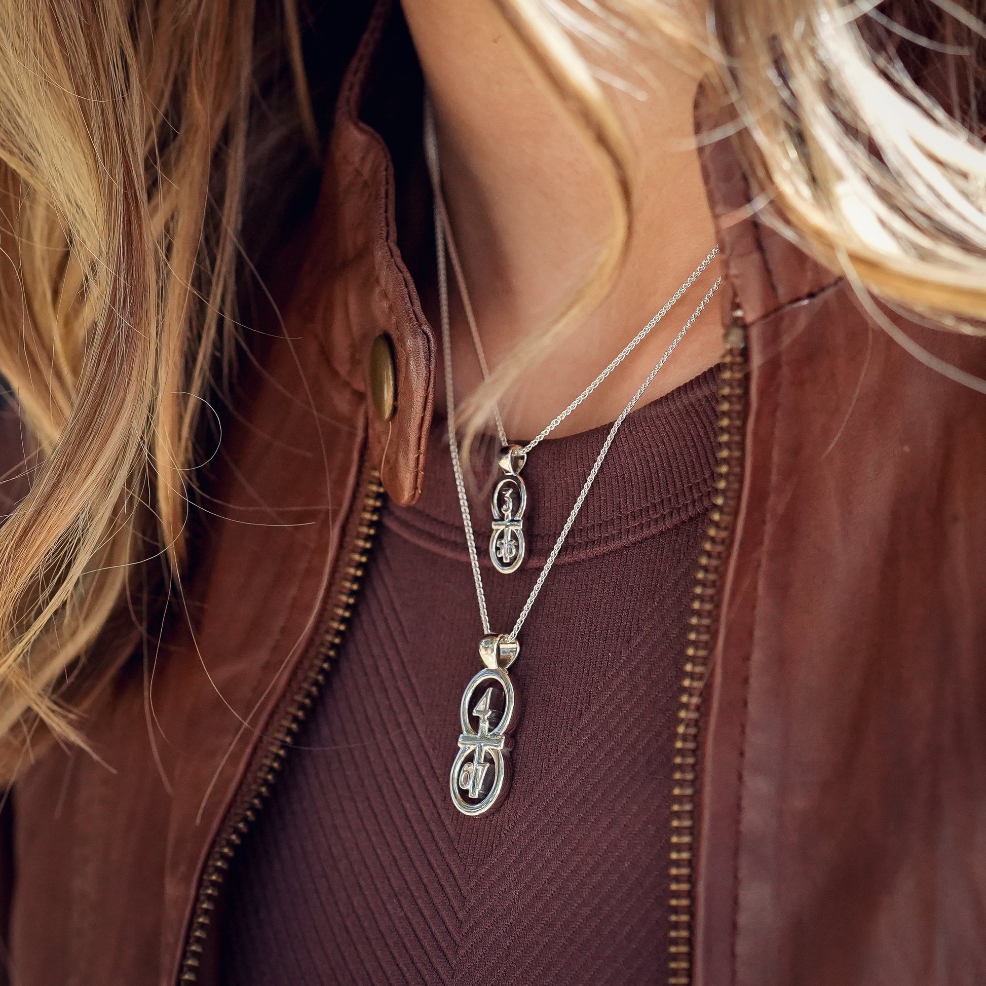 The silver RIYAN 29 and 11® pendant has the numbers 4,6, and 7 intertwined with the cross to represent Philippians 4:6-7 with the chapter name "Phillipians" inscribed on the back. Model is wearing a large silver Riyan pendant on our 18" Wheat chain and a small silver Riyan pendant on our 16" Wheat chain.  Model is wearing a burgundy tank and a brown leather jacket.