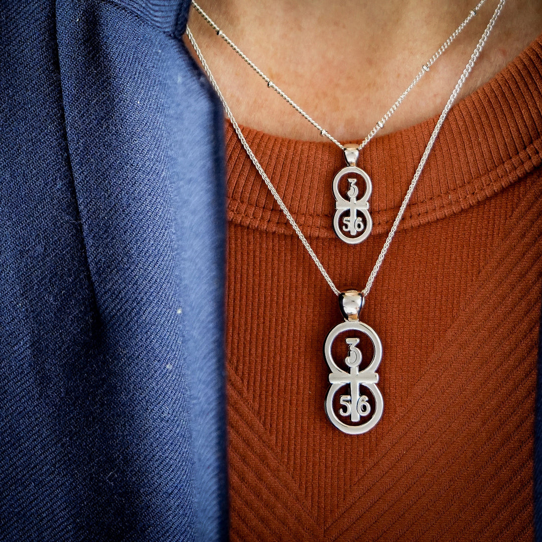 The RIYAN 29 and 11® silver pendant has the numbers 3, 5, and 6 intertwined with the cross to represent Proverbs 3:5-6 with the chapter word "Proverbs" inscribed on the back. Model is wearing our large silver Riyan pendant on an 18" Wheat chain and a small Riyan pendant on a 16" Satellite chain. Model is wearing a burnt orange tank and a Veronica Beard blazer.
