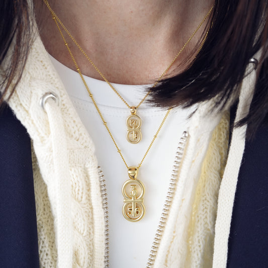The RIYAN 29 and 11® pendant has the numbers 3, 5, and 6 intertwined with the cross to represent Proverbs 3:5-6 with the chapter word "Proverbs" inscribed on the back.  The model is wearing our large Riyan pendant on our 18" satellite chain with our small Riyan pendant on our 16" Wheat chain.  Model is wearing a high neck white t-shirt, a navy Veronica Beard blazer, and cream sweater.