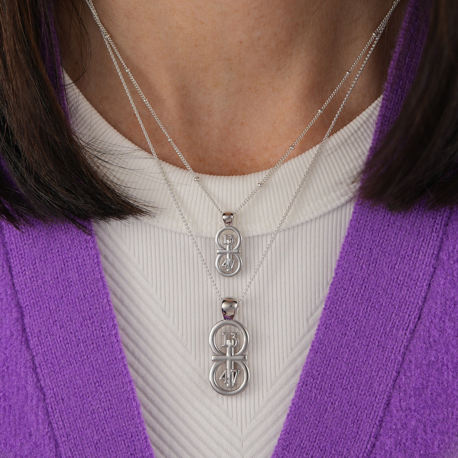 The silver RIYAN 29 and 11® pendant has the numbers 13, 4, and 4 intertwined with the cross to represent 1 Corinthians 13:4-7 with the chapter word "1 Corinthians" inscribed on the back.  Model is wearing the large silver pendant on an 18" Wheat chain and a small Riyan pendant on a 16" satellite chain.  Model is wearing a white high neck tank and purple sweater.