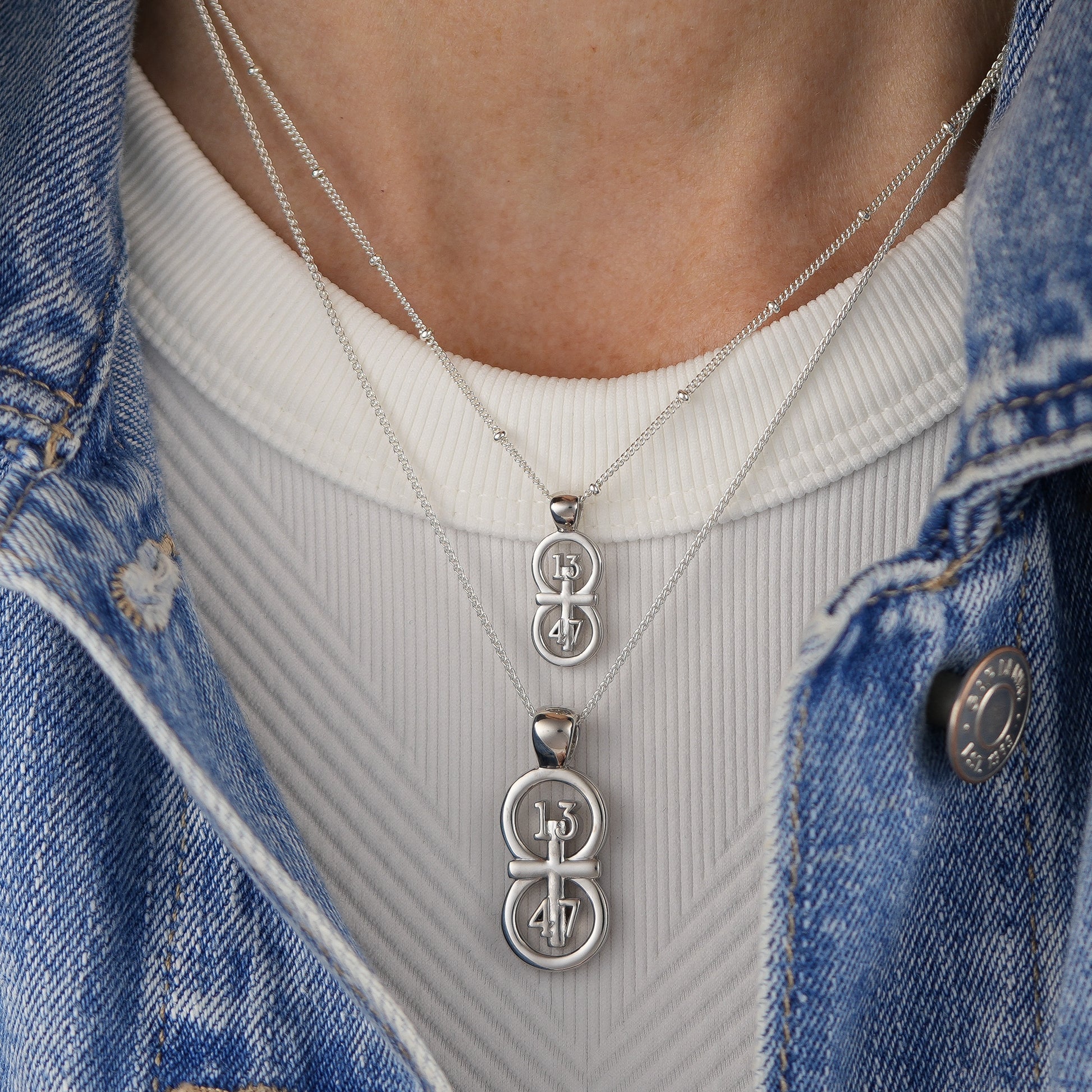 The silver RIYAN 29 and 11® pendant has the numbers 13, 4, and 4 intertwined with the cross to represent 1 Corinthians 13:4-7 with the chapter word "1 Corinthians" inscribed on the back.  Model is wearing the large silver pendant on an 18" Wheat chain and a small Riyan pendant on a 16" satellite chain.  Model is wearing a high neck white tank and denim jacket.