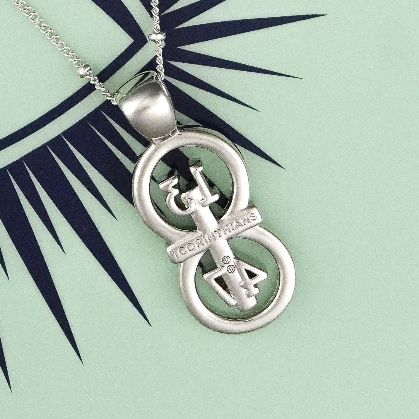 The silver RIYAN 29 and 11® pendant has the numbers 13, 4, and 4 intertwined with the cross to represent 1 Corinthians 13:4-7 with the chapter word "1 Corinthians" inscribed on the back. Pendant is facing backwards to show the chapter name.