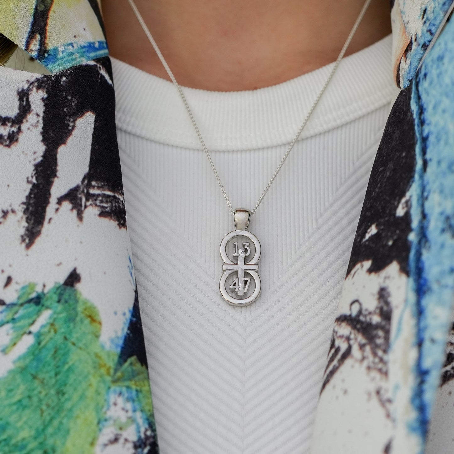 The silver RIYAN 29 and 11® pendant has the numbers 13, 4, and 4 intertwined with the cross to represent 1 Corinthians 13:4-7 with the chapter word "1 Corinthians" inscribed on the back.  Model is wearing the large silver pendant on an 18" Wheat chain.  Model is wearing a printed blazer in greens and blues.