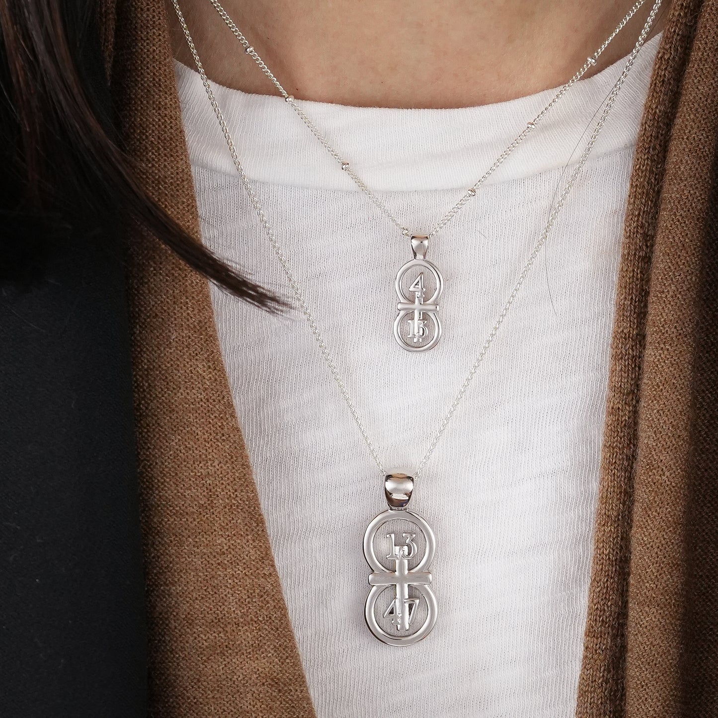 The silver RIYAN 29 and 11® pendant has the numbers 13, 4, and 4 intertwined with the cross to represent 1 Corinthians 13:4-7 with the chapter word "1 Corinthians" inscribed on the back.  Model is wearing the large silver pendant on an 18" Wheat chain and a small Riyan pendant on a 16" satellite chain.  Model is wearing a Veronica Beard blazer and brown scarf against a white tank.