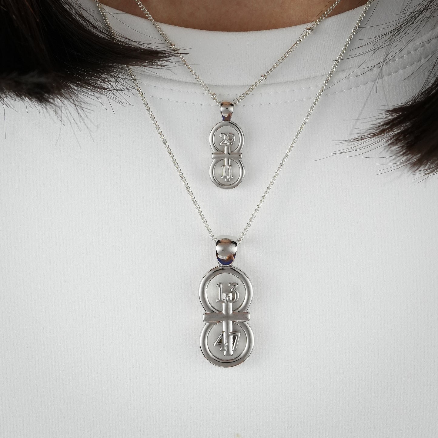 The silver RIYAN 29 and 11® pendant has the numbers 13, 4, and 4 intertwined with the cross to represent 1 Corinthians 13:4-7 with the chapter word "1 Corinthians" inscribed on the back.  Model is wearing the large silver pendant on an 18" Wheat chain and a small Riyan pendant on a 16" satellite chain.  Model is wearing a white high neck t-shirt.