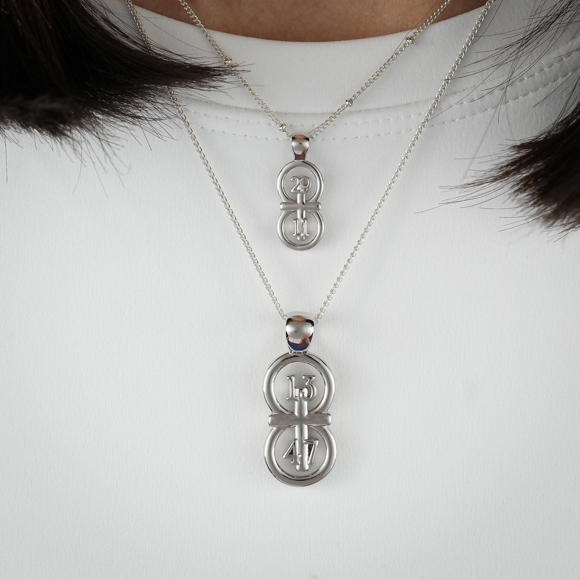 The silver RIYAN 29 and 11® pendant has the numbers 13, 4, and 4 intertwined with the cross to represent 1 Corinthians 13:4-7 with the chapter word "1 Corinthians" inscribed on the back.  Model is wearing the large silver pendant on an 18" Wheat chain and a small Riyan pendant on a 16" satellite chain.  Model is wearing a white high neck t-shirt.
