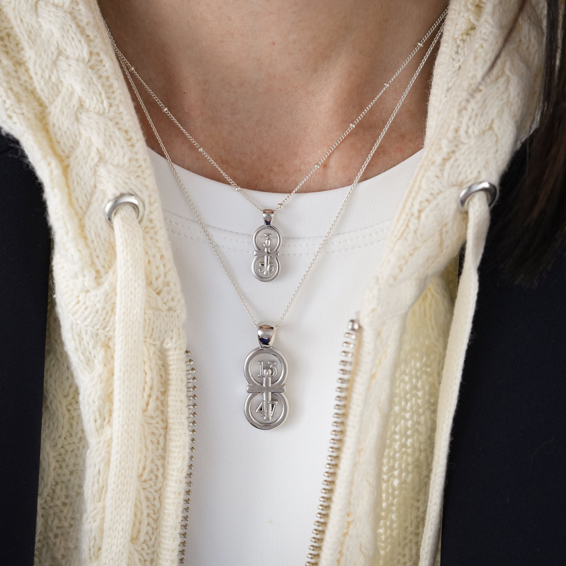 The silver RIYAN 29 and 11® pendant has the numbers 13, 4, and 4 intertwined with the cross to represent 1 Corinthians 13:4-7 with the chapter word "1 Corinthians" inscribed on the back.  Model is wearing the large silver pendant on an 18" Wheat chain and a small Riyan pendant on a 16" satellite chain.  Model is wearing a Veronica Beard blazer and cream sweater.
