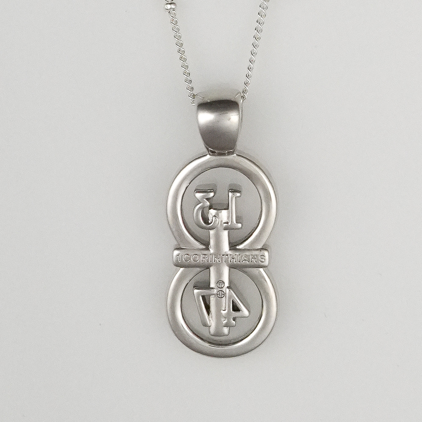 The silver RIYAN 29 and 11® pendant has the numbers 13, 4, and 4 intertwined with the cross to represent 1 Corinthians 13:4-7 with the chapter word "1 Corinthians" inscribed on the back.  Pendant is on a white background.