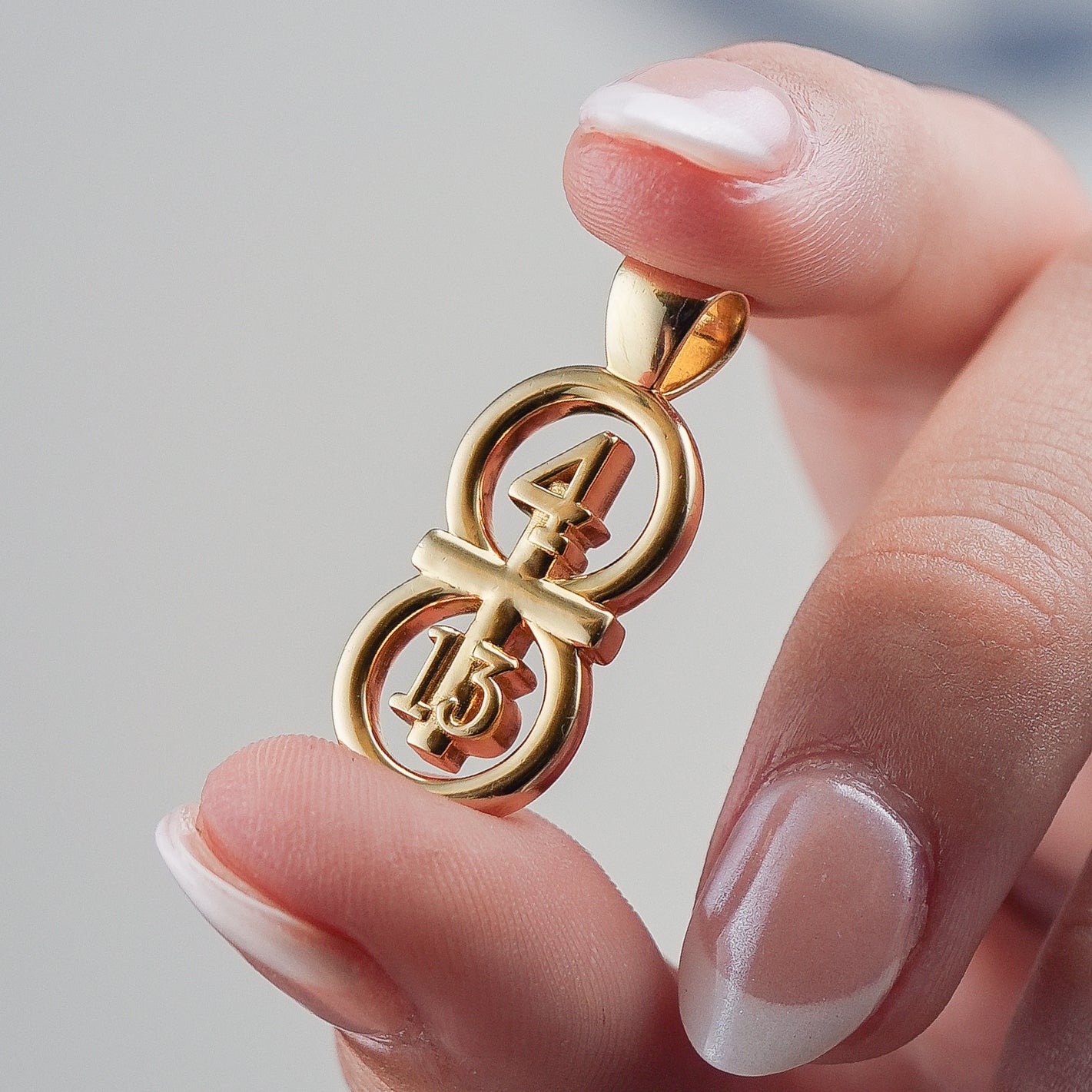 Our gold large pendant displayed on a white background held between two fingers for size reference. Our 29 and 11® pendant has the numbers 4 and 13 intertwined with the cross to represent Philippians 4:13 with the chapter word "Philippians" inscribed on the back.