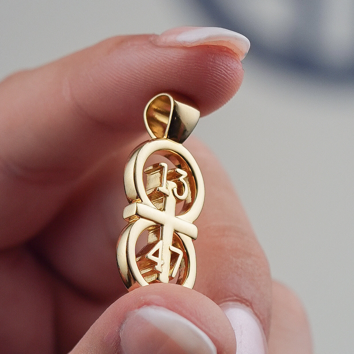 Closeup of our Gold pendant front size being held between two fingers for scale.  The RIYAN 29 and 11® pendant has the numbers 13, 4, and 4 intertwined with the cross to represent 1 Corinthians 13:4-7 with the chapter word "1 Corinthians" inscribed on the back.