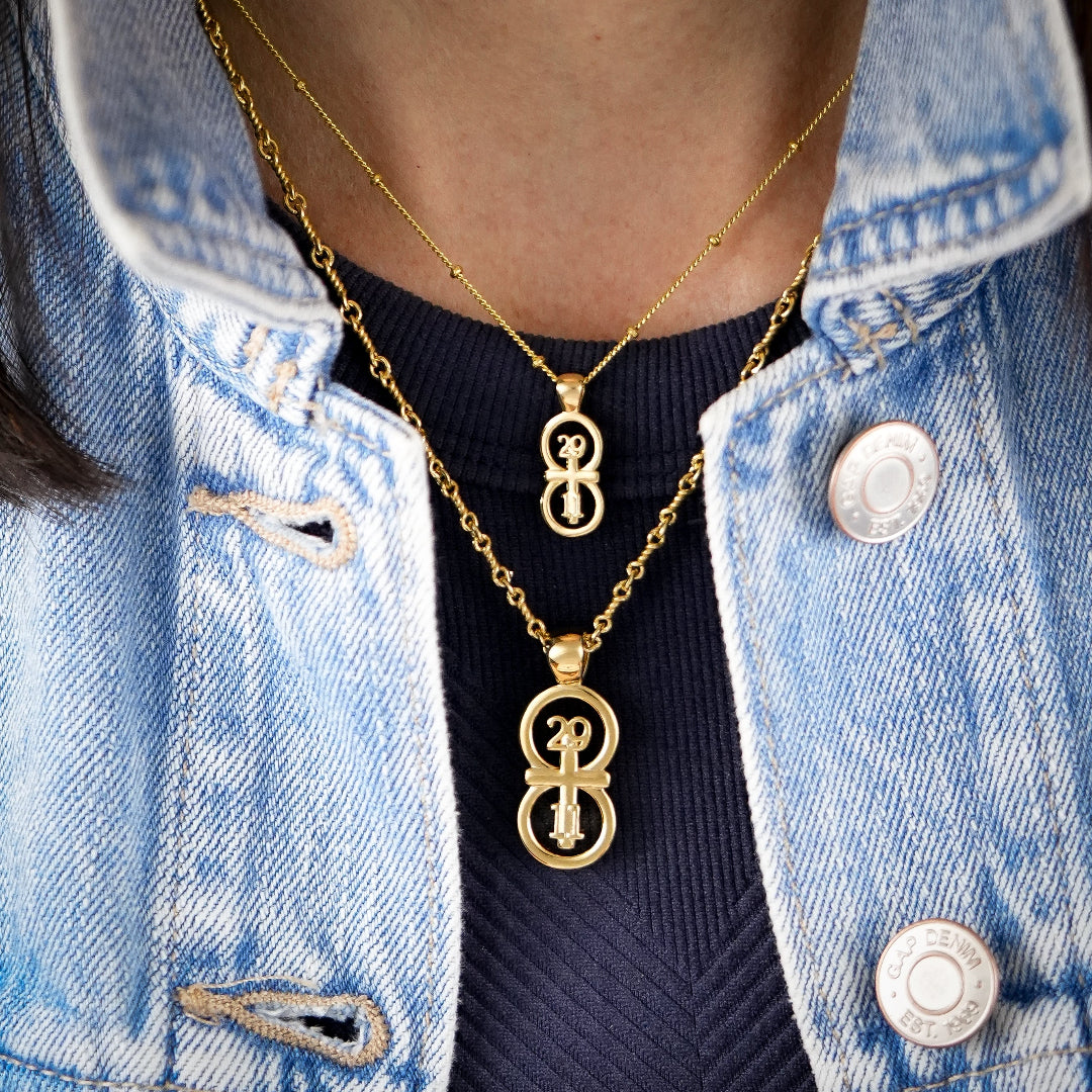The RIYAN 29 and 11® pendant has the numbers 29 and 11 intertwined with the cross to represent Jeremiah 29:11 with the chapter word "Jeremiah" inscribed on the back. n The model is wearing a large Riyan pendant on our 18" Rolo chain with a small Riyan pendant on our 16" Satellite chain.  Model is wearing a navy high neck tank and a denim jacket.