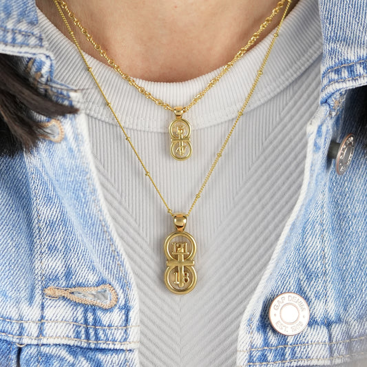 The RIYAN 29 and 11® pendant has the numbers 121, 1, and 8 intertwined with the cross to represent Psalm 121:1-8 with the chapter word "Psalm" inscribed on the back.  Model is wearing our large Riyan pendant on our 18" Satellite chain and our small Riyan pendant on our 16" Rolo chain.  Model is wearing a high neck white tank and denim jacket.