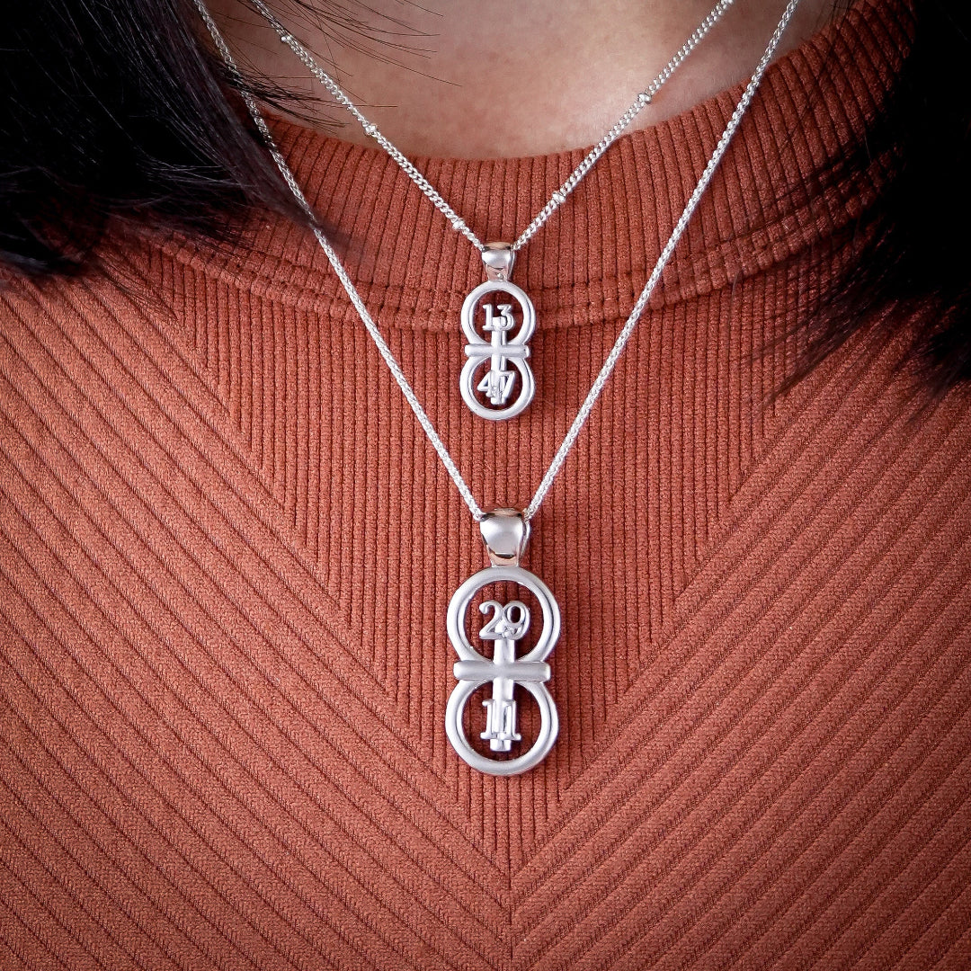 The RIYAN 29 and 11® silver pendant has the numbers 29 and 11 intertwined with the cross to represent Jeremiah 29:11 with the chapter word "Jeremiah" inscribed on the back.  Model is wearing the large silver Riyan pendant on an 18" Wheat chain and a small silver Riyan pendant on a 16" Satellite chain.  Model is wearing a high neck white burnt orange tank.