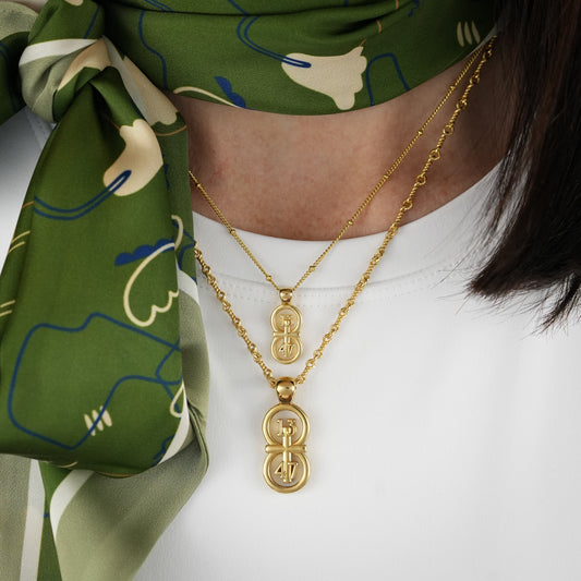 The RIYAN 29 and 11® pendant has the numbers 13, 4, and 4 intertwined with the cross to represent 1 Corinthians 13:4-7 with the chapter word "1 Corinthians" inscribed on the back. The model is wearing a large Riyan pendant on our 18" Rolo Chain and a small Riyan pendant on our 16" Satellite chain.  Model is wearing a high neck white t-shirt with a green scarf.