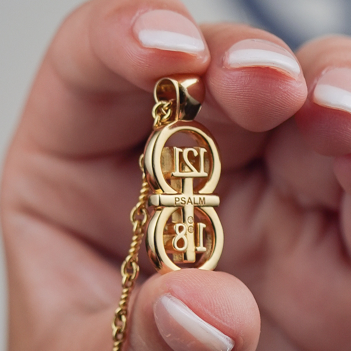 Our gold large pendant with the back displayed to show engraving detail on a white background held between two fingers for size reference.The RIYAN 29 and 11® pendant has the numbers 121, 1, and 8 intertwined with the cross to represent Psalm 121:1-8 with the chapter word "Psalm" inscribed on the back.