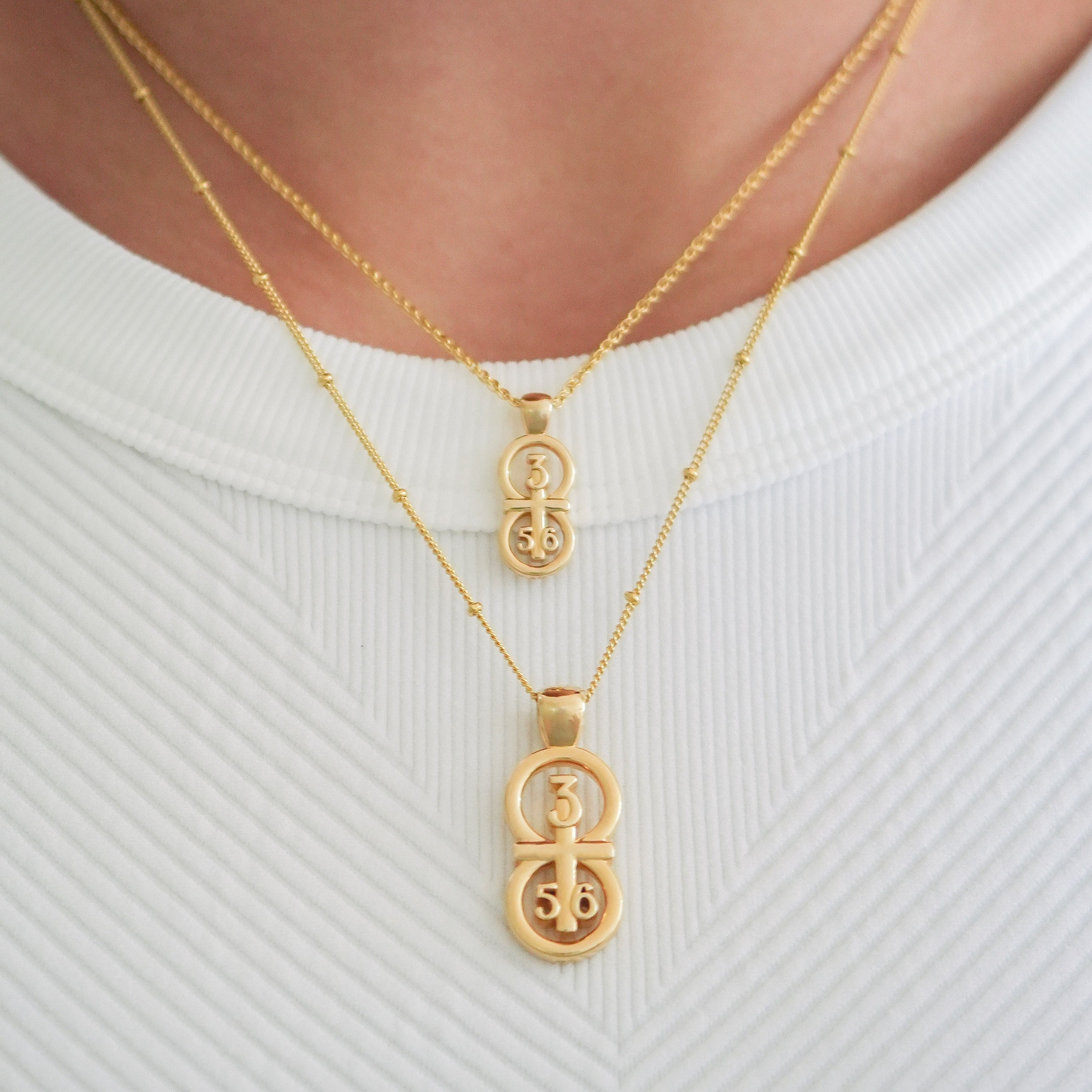 The RIYAN 29 and 11® pendant has the numbers 3, 5, and 6 intertwined with the cross to represent Proverbs 3:5-6 with the chapter word "Proverbs" inscribed on the back.  Model is wearing both size pendants.  The small is on our 16" wheat chain and the large is on our 18" satellite chain.  Model is wearing a white ribbed Free People Movemint tank.