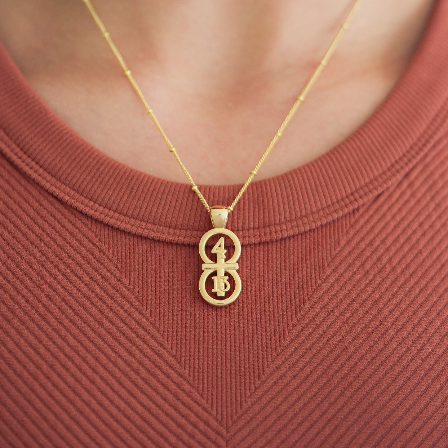 Gold large pendants displayed on the neck against a burnt orange shirt with our 14k gold filled satellite chain.  Our 29 and 11® pendant has the numbers 4 and 13 intertwined with the cross to represent Philippians 4:13 with the chapter word "Philippians" inscribed on the back.